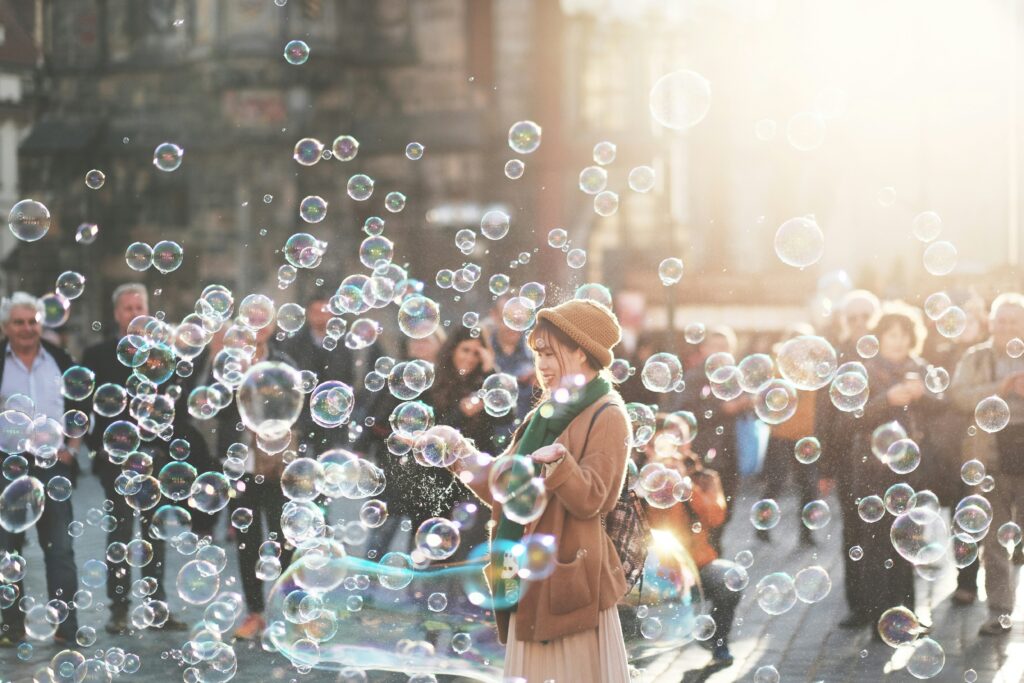A young woman blowing bubbles while standing in a crowd of people in a public outdoor space.