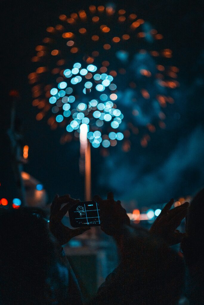 A person holding up a phone to capture fireworks lighting up the night sky.
