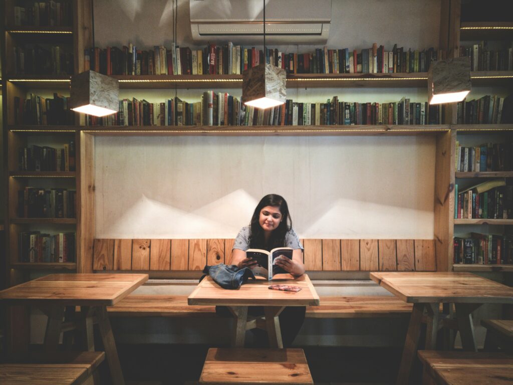 A young woman sitting alone in a library, reading a book at a table surrounded by shelves.